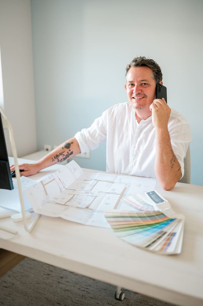 Inhaber Markus sitzt am Schreibtisch in Gleisdorf mit einem Farbfächer für Wandfarben und telefoniert.